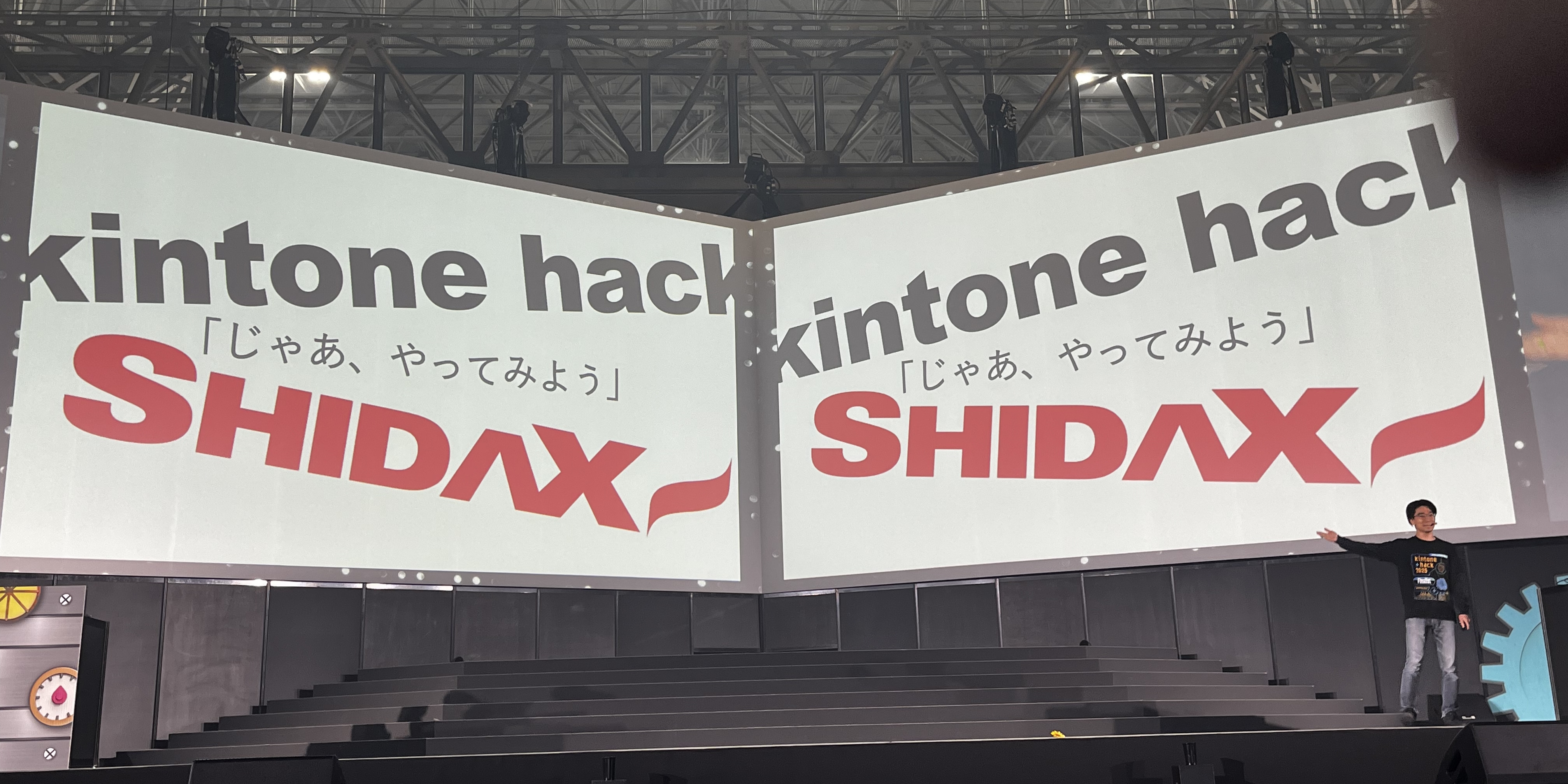 kintone hack 2025 本戦の最後のワンシーン。「kintone hack」「SHIDAX」という大きな文字と、「じゃあ、やってみよう」というやや小さな文字が巨大なスクリーンいっぱいに映し出されており、その手前で志太がスライドを指している。